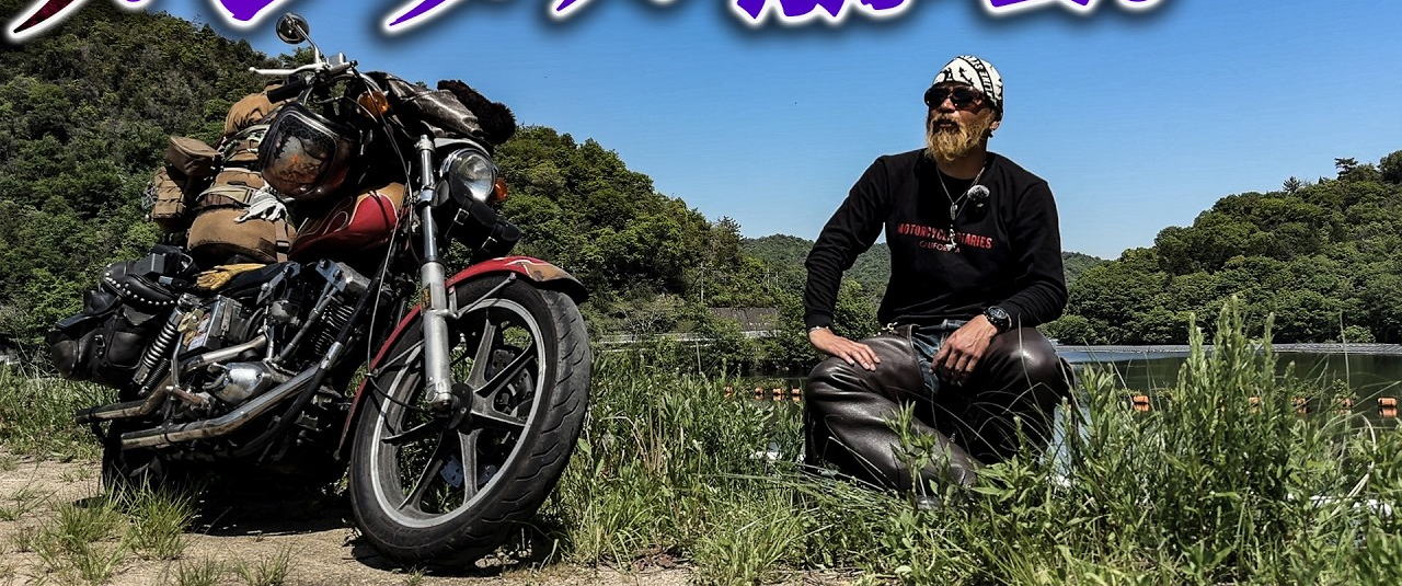 ミショメンTV の動画より。積載したバイクの脇、草地に座る人物と対岸の山